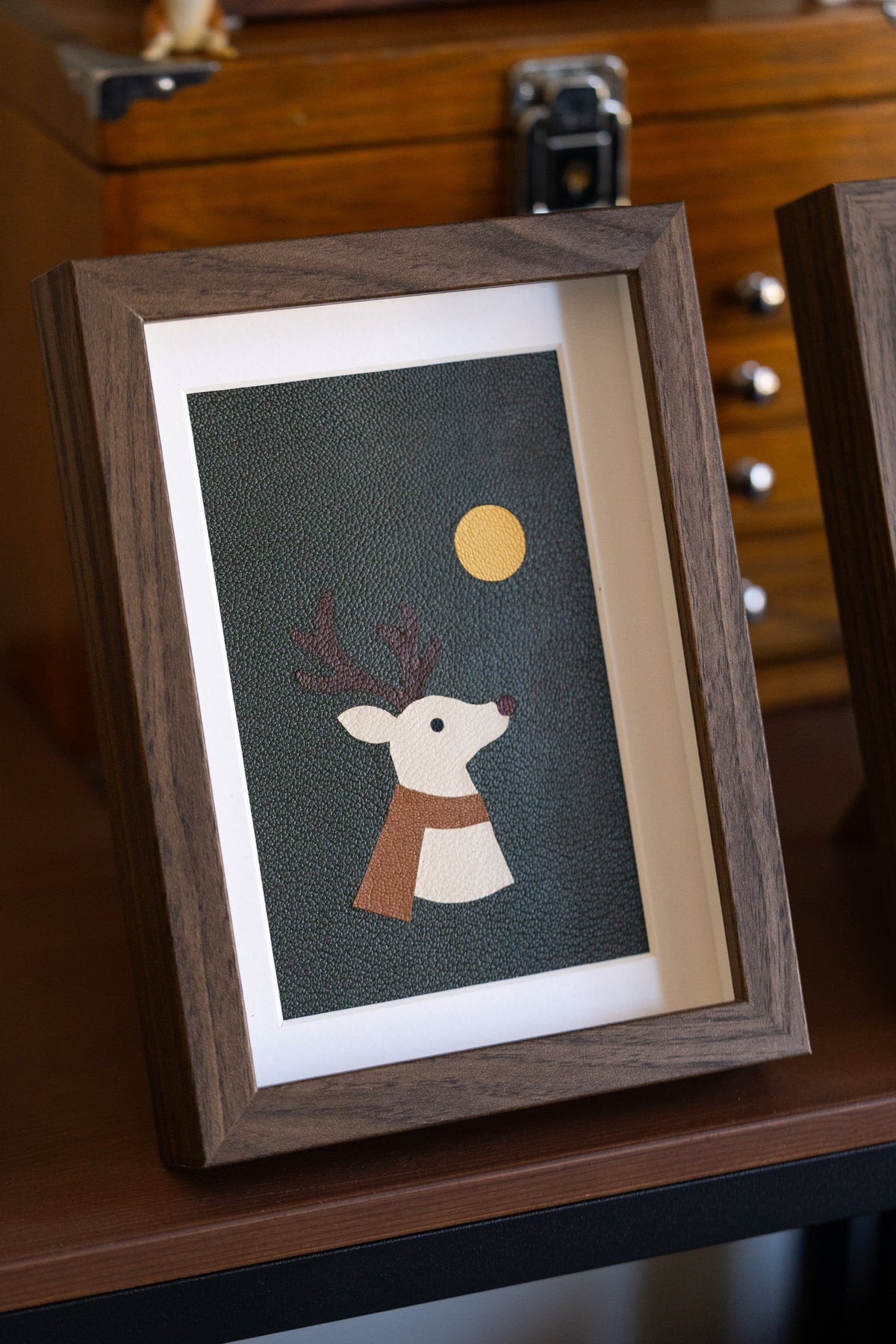 Reindeer leather art in a walnut frame, a cream reindeer with a rust scarf and yellow moon on green leather