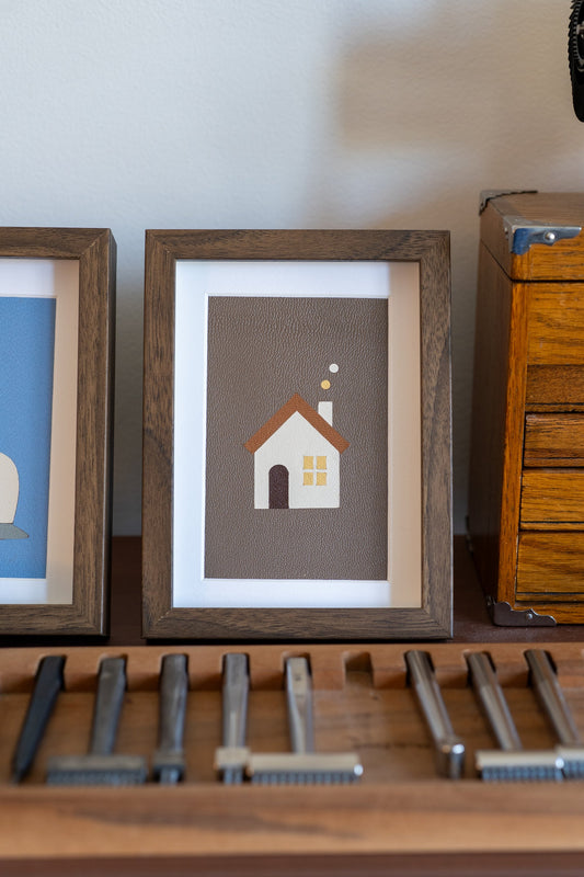 Warm Home leather art in a walnut frame, a white house with a brown roof on taupe leather